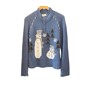 May include: A blue knit quarter-zip sweater featuring a winter scene with two white snowmen wearing hats and scarves, dark brown pine trees, and white snowflakes. The sweater has a ribbed collar, cuffs, and hem with white decorative stitching.