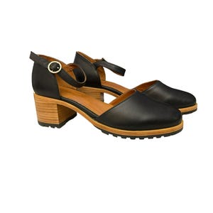Puede incluir: Sandalias de cuero negro con tacón, correa de tobillo con hebilla y plataforma de madera.