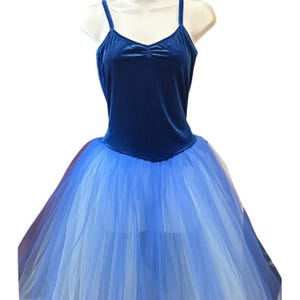 Może przedstawiać: Niebieski trykot i tutu baletowe. Góra wykonana jest z głębokiego, niebieskiego aksamitu ze spaghetti i dekoltem w kształcie serca. Tutu to warstwowy gradient od jasnego do średniego niebieskiego tiulu, tworzący pełną, płynącą spódnicę. Kostium jest wystawiony na manekinie.