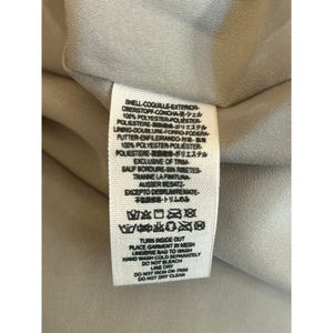 May include: A white fabric care label with black text detailing the fabric composition and care instructions. The label reads "SHELL-COQUILLE-EXTERIOR OBERSTOFF-CONCHA・シェル 100% POLYESTER-POLIESTER- POLIESTERE・ポリエステル LINING-DOUBLURE-FORRO-FODERA FUTTER-ENFLERANDO- 100% POLYESTER-POLIESTER- POLIESTERE-ポリエステル EXCLUSIVE OF TRIM SAUF BORDURE-SIN RABETES TRANNE LA FINITURA- AUSSER BESATZ EXCEPTO DEBRUMREMATE- 不振・トリムのみ TURN INSIDE OUT PLACE GARMENT IN MESH LINGENE BAG TO WASH HAND WASH COLD SEPURATELY DO NOT BLEACH LINE DRY DO NOT SRON ON TRIM DO NOT DRY CLEAN"