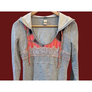 Peut inclure: Sweat-shirt &agrave; capuche gris avec un motif patchwork color&eacute; sur la poitrine. Le patchwork comprend des nuances de rose, d'orange et de noir. Le sweat-shirt a des manches longues et une poche avant.