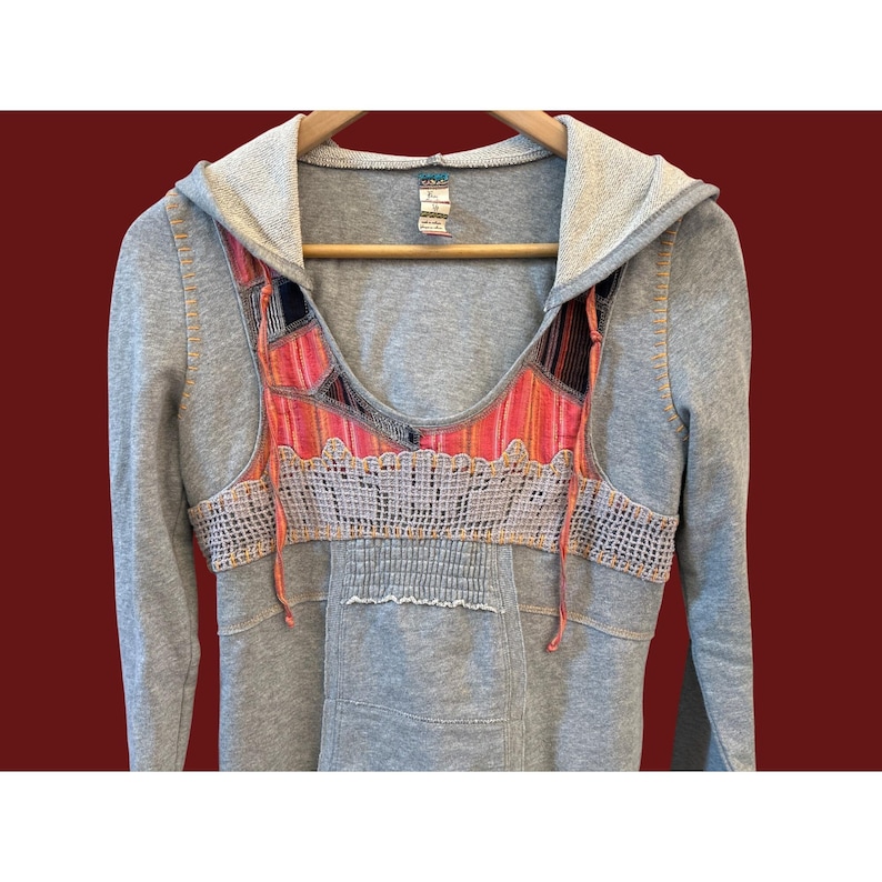 Peut inclure: Sweat-shirt &agrave; capuche gris avec un motif patchwork color&eacute; sur la poitrine. Le patchwork comprend des nuances de rose, d'orange et de noir. Le sweat-shirt a des manches longues et une poche avant.