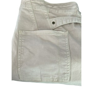 May include: A pair of beige cargo pants with a large patch pocket on the right side. The pocket has a stitched edge and is sewn onto the pants.