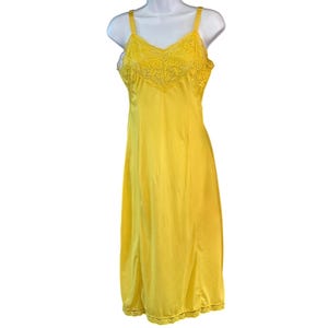 Könnte beinhalten: Ein leuchtend gelbes Unterkleid mit V-Ausschnitt und aufwendigen floralen Spitzendetails am Oberteil und an den Trägern. Das Kleid hat einen glatten, einfarbigen Rock, der bis zum Knie reicht und mit einer passenden Spitzenborte am Saum abschließt.