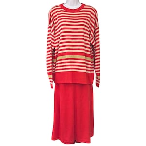 May include: A two-piece knit set consisting of a long-sleeved sweater and a matching skirt. The sweater features horizontal red and cream stripes with a single gold metallic stripe. The skirt is a solid red color with a simple, straight silhouette.
