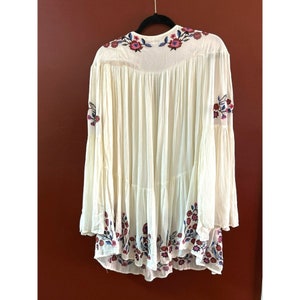May include: A white, embroidered, long-sleeved tunic with a tiered skirt and floral embroidery in shades of pink, blue, and green.