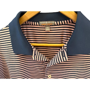 May include: A pink and navy blue striped polo shirt with a navy blue collar. The shirt is hanging on a wooden hanger. The brand name "PETER MILLAR" is visible on the collar.
