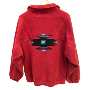 Könnte beinhalten: Rote Fleecejacke mit farbenfrohem geometrischem Design auf dem Rücken. Die Jacke hat einen hohen Kragen und lange Ärmel. Das Design hat schwarze, türkisfarbene, lila und gelbe Akzente.