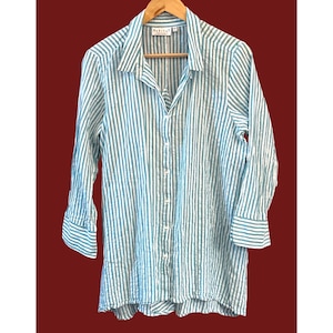 May include: A long-sleeved, button-down shirt with vertical stripes in teal and white. The shirt is hanging on a wooden hanger against a dark red background. The shirt has a collar and buttoned cuffs. The brand name "Habitat" is visible on the label.