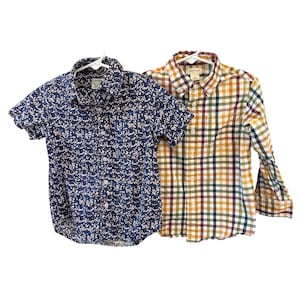 Puede incluir: Dos camisas abotonadas en perchas blancas. Una es de manga corta, azul marino con un estampado floral blanco. La otra es de manga larga, con un estampado de cuadros en tonos amarillos, verdes, marrones y blancos.