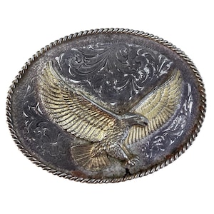 May include: An oval belt buckle featuring a gold-toned eagle with outstretched wings centred on a dark, silver-toned background with intricate floral scrollwork engravings. The edge of the buckle is finished with a twisted rope-style border.