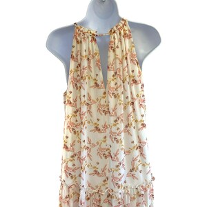 Pode incluir: Um vestido maxi branco sem mangas com estampa floral em tons de rosa e amarelo. O vestido tem um decote nas costas em forma de fechadura e uma bainha com babados.