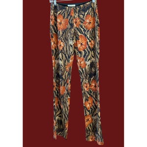 Puede incluir: Un pantalón estampado con un diseño floral. El pantalón presenta una cintura negra y un estampado de flores naranjas y un estampado similar a la piel de serpiente en tonos marrones y dorados. Cuelga de una percha.