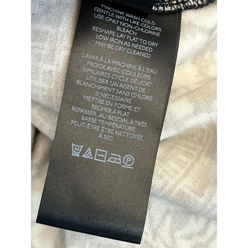 May include: A black clothing tag with white text detailing washing instructions. The tag includes information on machine washing cold, using non-chlorine bleach, reshaping, and low-temperature ironing. The tag also has a series of laundry symbols.