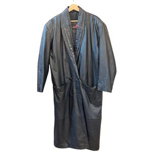 May include: A long, black leather coat with a double-breasted design. The coat features a notched collar, two front pockets, and a pleated detail on the lapel. The coat is hanging on a wooden hanger.