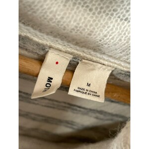 May include: Close-up of a garment tag with the brand name "Moth" and size "M". The tag also indicates the item was made in China. The garment appears to be a sweater or cardigan, with a striped pattern.