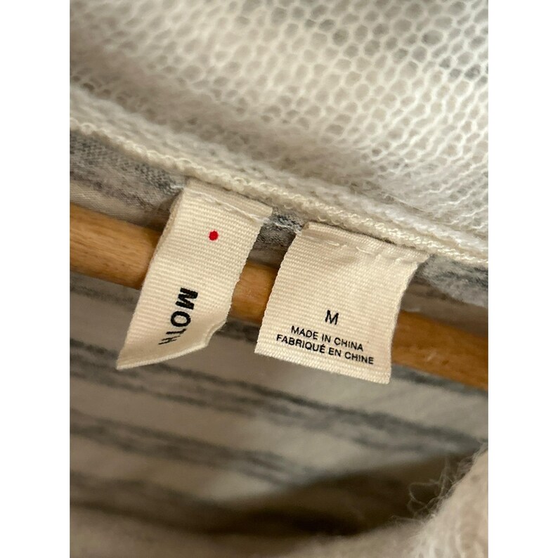 May include: Close-up of a garment tag with the brand name "Moth" and size "M". The tag also indicates the item was made in China. The garment appears to be a sweater or cardigan, with a striped pattern.