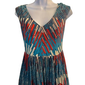 Op de afbeelding: Een mouwloze jurk met een V-hals en een geometrisch patroon in blauw, wit en rood. De jurk heeft een geplooide rok.