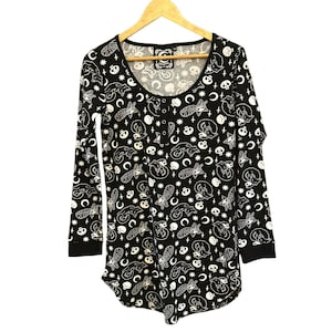 May include: Black long-sleeved shirt with a white print of cats, moons, stars, and skulls. The shirt has a scoop neck and button closure.