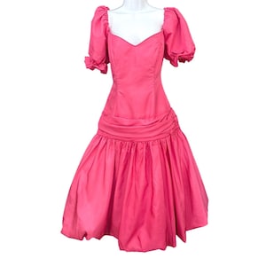 May include: A vibrant hot pink dress featuring short puff sleeves with gathered cuffs, a sweetheart neckline, and a fitted bodice. The waistline is defined by a horizontal gathered band, transitioning into a voluminous, pleated bubble-style skirt.