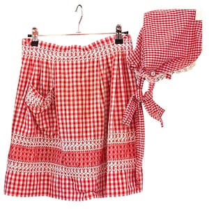 Pode incluir: Avental e touca xadrez vermelha e branca a condizer. O avental tem um bolso e bordados decorativos. A touca tem uma renda e laços. O conjunto está pendurado num cabide de metal.