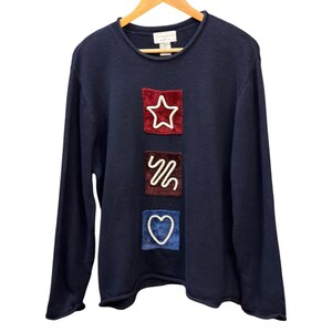 Könnte beinhalten: Ein dunkelblauer Langarmpullover mit drei übereinander angeordneten quadratischen Aufnähern in Rot, Dunkelbraun und Blau. Jeder Aufnäher zeigt eine weiße gestickte Kontur in Form eines Sterns, einer Wellenlinie und eines Herzens.
