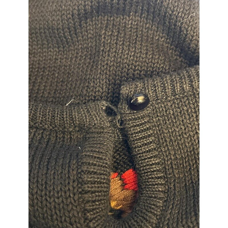 May include: Close-up of a dark gray knit cardigan with a single black button. The button is fastened, and the fabric is slightly stretched. A small patch of red and brown embroidery is visible near the button closure.