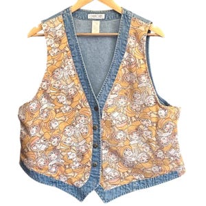 Könnte beinhalten: Eine Jeansweste mit V-Ausschnitt und Knopfverschluss. Die Weste hat eine hellblaue Jeansaußenseite mit einem gemusterten Innenfutter. Das Innenmuster ist ein sich wiederholendes Design von stilisierten Gesichtern in Gelb-, Braun- und Weißtönen. Der Markenname "Cherokee" ist sichtbar.