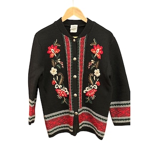 Op de afbeelding: Zwarte gebreide cardigan met rode, witte en groene geborduurde bloemenmotieven. De cardigan heeft een knoopsluiting en een geribbelde breistructuur.
