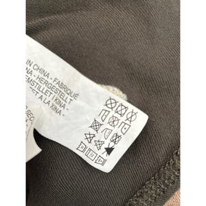 May include: Close-up of a white clothing tag with black symbols and text. The tag is partially visible, showing care instructions and phrases in multiple languages, including "IN CHINA - FABRIQU&Eacute;". The tag is on a dark olive green fabric.