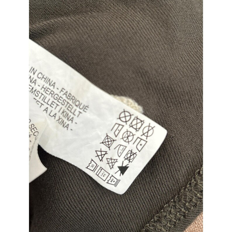 May include: Close-up of a white clothing tag with black symbols and text. The tag is partially visible, showing care instructions and phrases in multiple languages, including "IN CHINA - FABRIQU&Eacute;". The tag is on a dark olive green fabric.