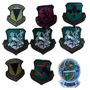 May include: A collection of embroidered military patches in various shield shapes. The patches feature diverse designs, including a dragon, a deer-drawn chariot, and geometric shapes, with text such as "Virtus Perdurat" and "Strategic Air Command".
