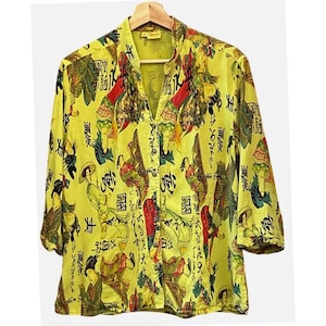 May include: Yellow button-down shirt with a mandarin collar and three-quarter sleeves. The shirt features a repeating pattern of figures in traditional dress and calligraphy in black, red, and green. The shirt is hanging on a wooden hanger.