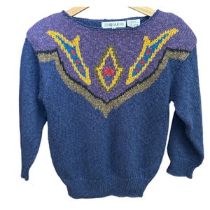 May include: A blue knit sweater with a decorative design on the upper chest. The design features a purple background with gold, red, and blue accents. The sweater has long sleeves and a round neckline. The brand name "SEGUE" is visible on the label.