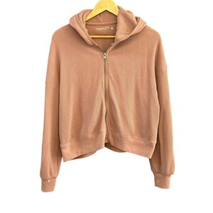 May include: A light pink waffle knit zip-up hoodie with a hood and long sleeves. The hoodie has a cropped fit and a zipper closure.