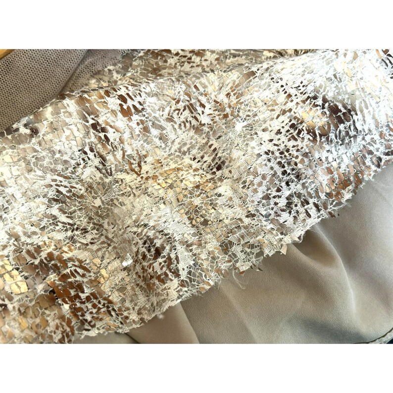 May include: Close-up of a white lace fabric with gold accents. The lace has a delicate pattern and is soft and flowing.