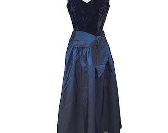 May include: A sleeveless dress featuring a dark navy blue velvet bodice with a sweetheart neckline and a contrasting iridescent blue taffeta skirt. The waist is accented with a wide, gathered fabric sash that ties into a bow on the side.