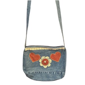 May include: A denim crossbody bag with a decorative design. The bag features two red plaid hearts and a cream-colored crocheted flower with a rhinestone center. The top edge is trimmed with lace.