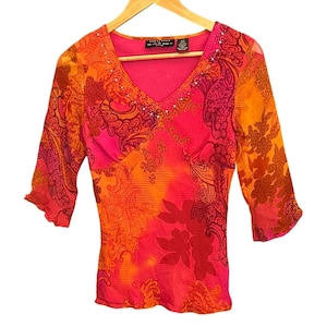 Könnte beinhalten: Ein lebhaftes, gemustertes Oberteil in Pink-, Orange- und Rottönen. Die Bluse mit V-Ausschnitt hat Dreiviertelärmel und ist mit kleinen, glitzernden Verzierungen am Ausschnitt versehen. Der Stoff hat einen transparenten Überzug mit Blumen- und Paisley-Design.