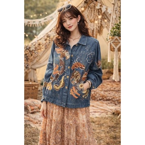 May include: A denim jacket with colorful embroidered designs. The jacket features a button-up front, long sleeves, and pockets. The embroidery includes orange, brown, and yellow patterns. The jacket is worn over a long, patterned skirt.