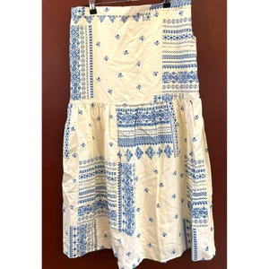 Op de afbeelding: Crèmekleurige rok met een blauw patchwork ontwerp. De rok heeft een hoge taille en een uitlopende zoom van middellange lengte. De blauwe patronen omvatten bloemen- en geometrische motieven, wat een vintage esthetiek creëert. De rok hangt aan een hanger.