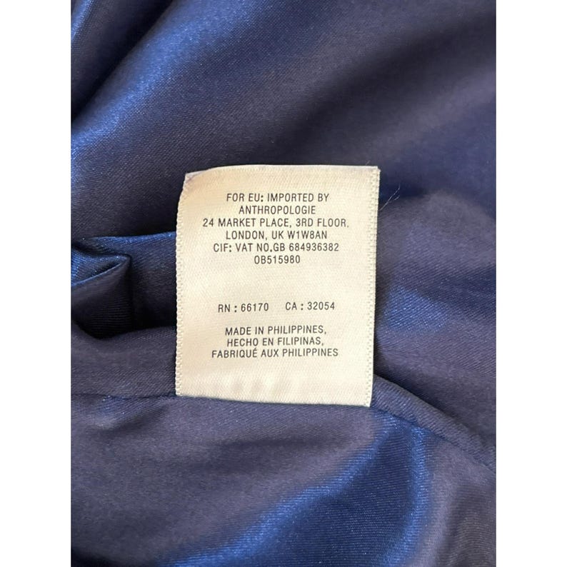 Puede incluir: Una etiqueta blanca con texto negro unida a una tela azul oscuro. La etiqueta contiene informaci&oacute;n sobre el fabricante, Anthropologie, y el origen del producto, que est&aacute; hecho en Filipinas.