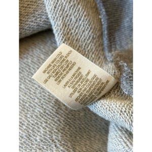 Peut inclure: Gros plan d'un sweat-shirt gris avec une &eacute;tiquette blanche. L'&eacute;tiquette contient des instructions d'entretien en anglais et en fran&ccedil;ais, notamment "Machine wash cold" et "Tumble dry low". Le tissu semble &ecirc;tre un tricot doux et textur&eacute;.