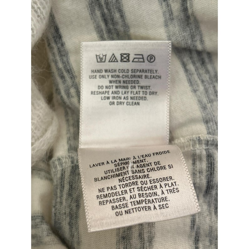 May include: Close-up of a white and gray striped fabric with two care instruction labels attached. The labels provide washing and drying instructions, including hand washing, non-chlorine bleach, and low iron.