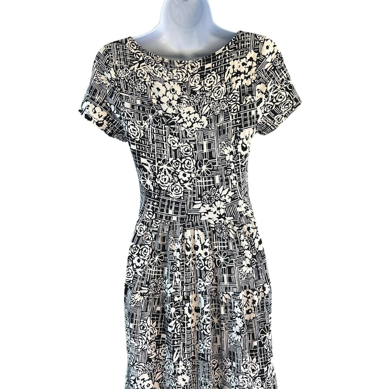 May include: A black and white patterned dress with a floral and geometric design. The dress features short sleeves and a fitted bodice that flows into a pleated skirt. The pattern includes roses and grid-like shapes.