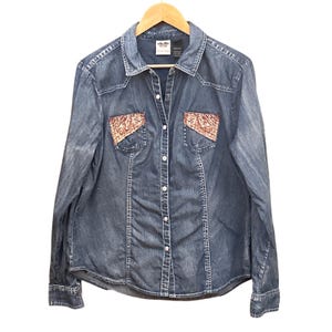 Może przedstawiać: Jeansowa koszula zapinana na guziki z kołnierzykiem i długimi rękawami. Koszula posiada ozdobne, haftowane naszywki na kieszeniach na piersi i małą metkę z logo. Prezentowana jest na drewnianym wieszaku.