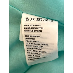 May include: White care label with black text detailing the fabric composition and care instructions for a garment. The label reads: "MAIN: 100% RAMIE LINING: 100% COTTON EXCLUSIVE OF TRIMS COLD HAND WASH SEPARATELY PULL INTO SHAPE DO NOT BLEACH OR WRING DO NOT TUMBLE DRY DRY FLAT IN SHADE WARM IRON IF REQUIRED DRY CLEANABLE (P) MADE IN CHINA DESIGNED IN AUSTRALIA 57567DB"