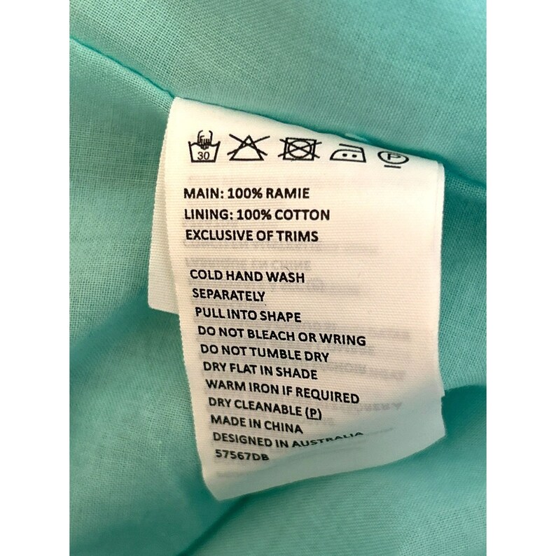 May include: White care label with black text detailing the fabric composition and care instructions for a garment. The label reads: "MAIN: 100% RAMIE LINING: 100% COTTON EXCLUSIVE OF TRIMS COLD HAND WASH SEPARATELY PULL INTO SHAPE DO NOT BLEACH OR WRING DO NOT TUMBLE DRY DRY FLAT IN SHADE WARM IRON IF REQUIRED DRY CLEANABLE (P) MADE IN CHINA DESIGNED IN AUSTRALIA 57567DB"