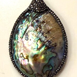 May include: A pear-shaped pendant with a shimmering abalone shell center, framed by a dark, beaded border. The shell displays iridescent colors of green, blue, and gold. A small, beaded cap tops the pendant.
