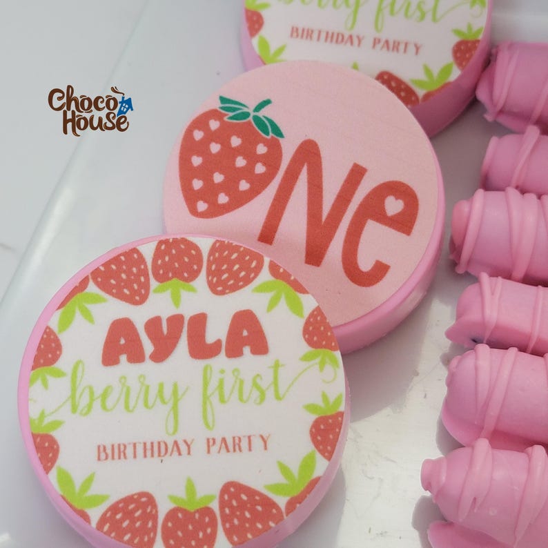 May include: Pink and white birthday party treats with strawberry designs. One treat has a large strawberry and the word "ONE". Another says "AYLA berry first BIRTHDAY PARTY". The treats are round and surrounded by pink chocolate-covered sticks.